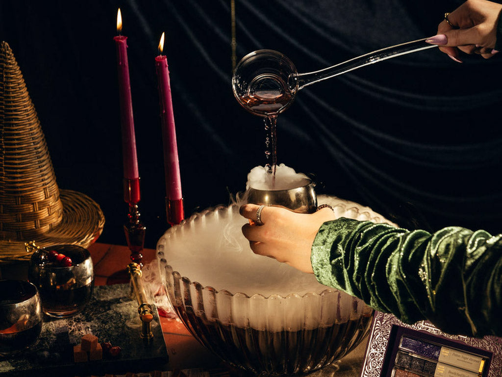 A person wearing a green velvet sleeve pours a clear liquid from a glass ladle into a large glass bowl filled with white smoke, set against a dark draped backdrop with lit pink candles and mystical props.