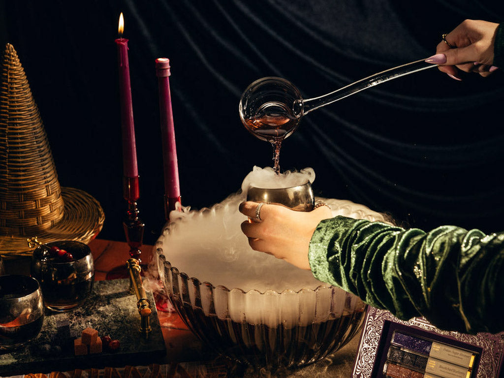 A person in a green velvet sleeve pours liquid from a glass ladle into a metal cup over a large bowl filled with dry ice and smoke, with lit pink candles, a woven witches hat, and dark draped velvet fabric in the background.