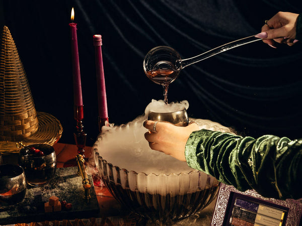 A person in a green velvet sleeve pours liquid from a glass ladle into a metal cup over a large bowl filled with dry ice and smoke, with lit pink candles, a woven witches hat, and dark draped velvet fabric in the background.