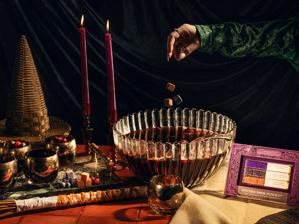 A styled Halloween-themed scene with a large ribbed glass punch bowl filled with dark red drink, surrounded by candles, metallic cups,cocktail mixer cubes, a witch hat, and a package labeled 'Witches Brew Punch' on a draped table against a dark curtain. A hand in a green sleeve drops cubes into the bowl.