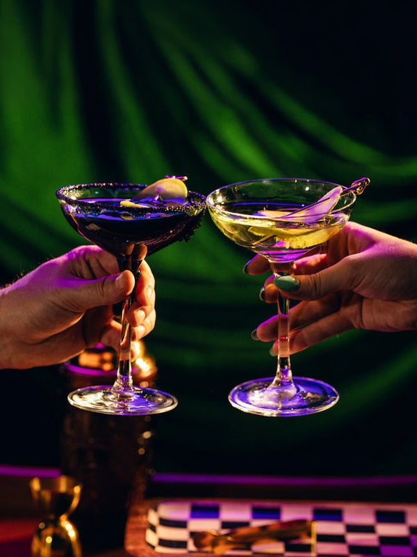 Two hands holding up cocktails in coupe glasses for a toast, one with a dark purple drink and a lime wheel, and one with a yellow drink and a lemon twist, against a dark green draped background.