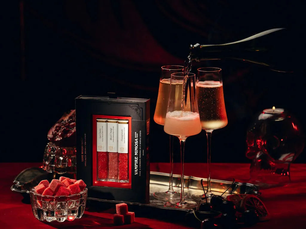 A boxed set of Vampire Windows red mood lighting candles sits on a red tablecloth next to three filled champagne flutes with pink liquid, surrounded by gothic props including a skull-shaped glass and a bowl of red wax cubes.