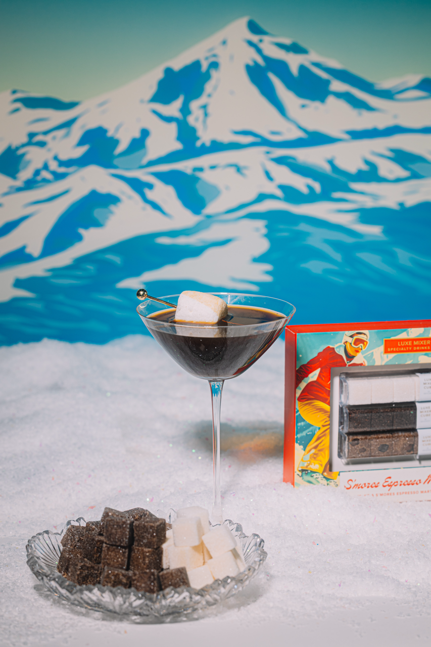 S'mores Esprersso Martini with marshmallow in a glass against a mountainous background, on a snowy surface.
