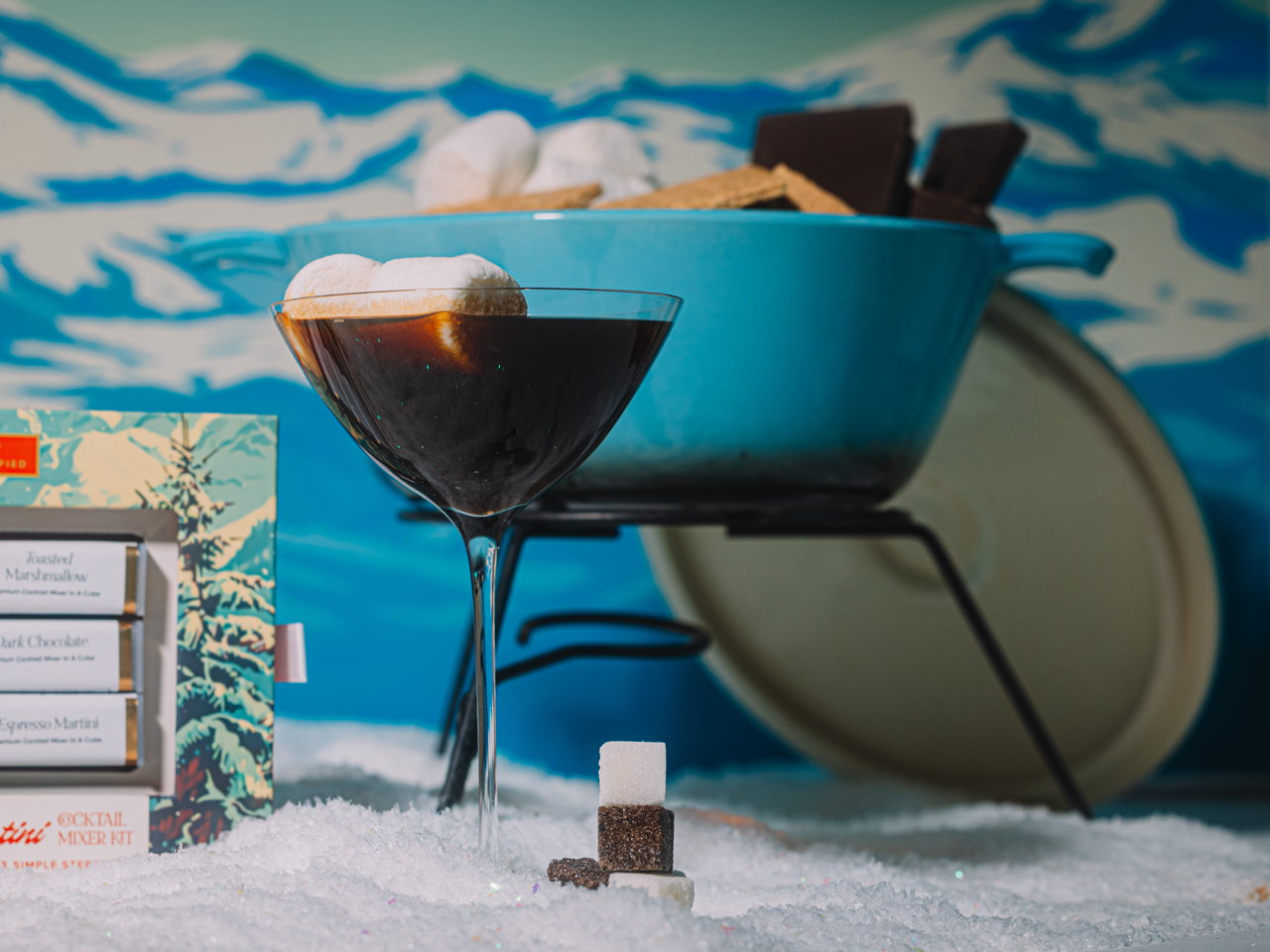 S'mores espresso martini with marshmallows in a snow-like setting with a blue bowl with S'mores components in the background.