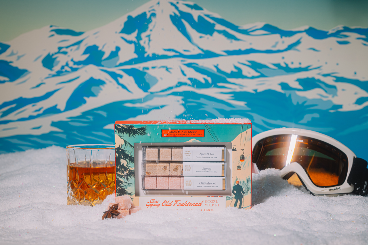 Snowy mountain landscape with a box of Chai Eggnog Old Fashioned cocktails, a glass of cocktail, and goggles.