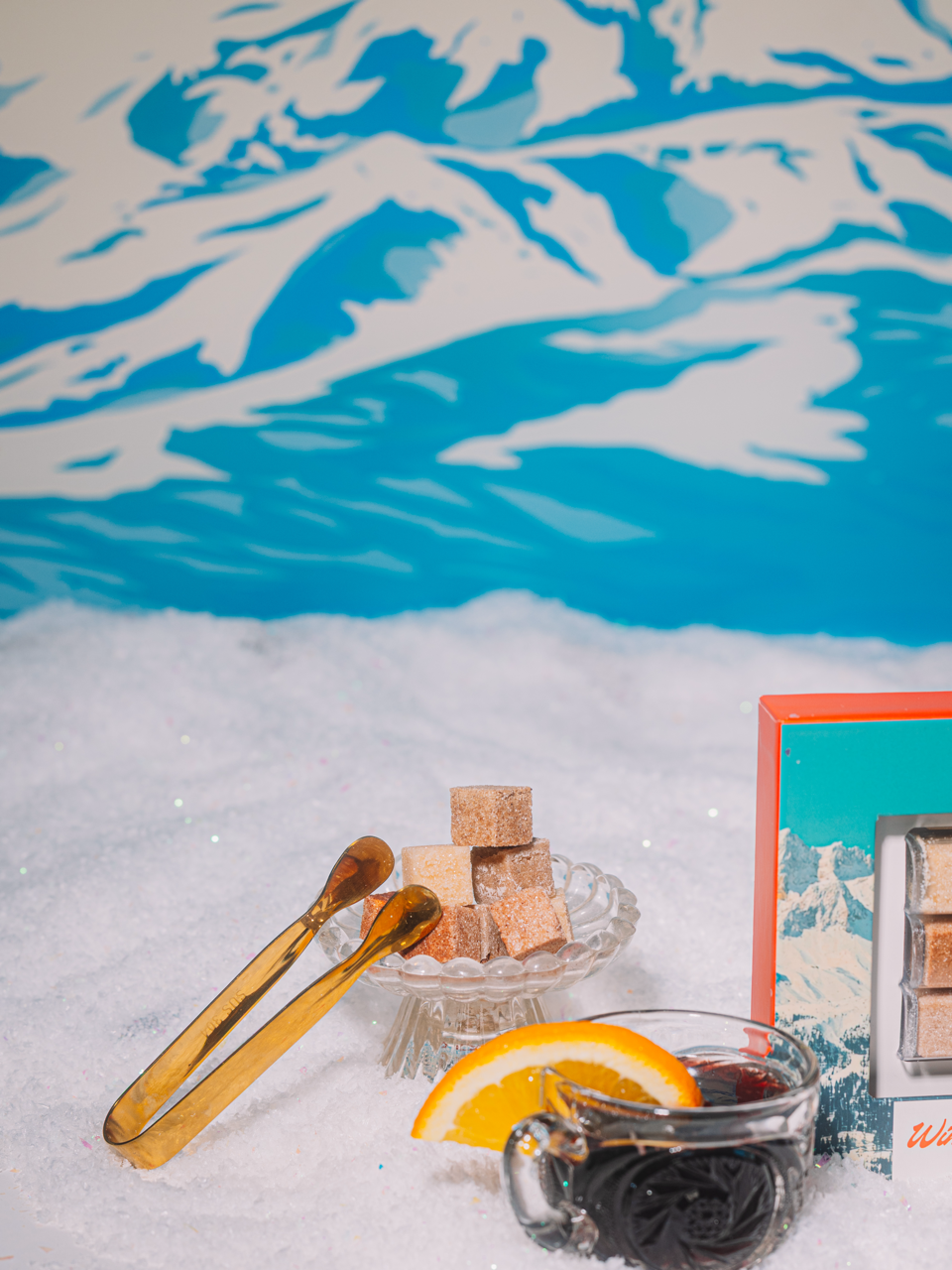 Sliced orange with a glass of mulled red wine on a snowy surface and a decorative plate with brown Luxe Mixer Cubes and Mini tongs. On a blue winter painting in the background.