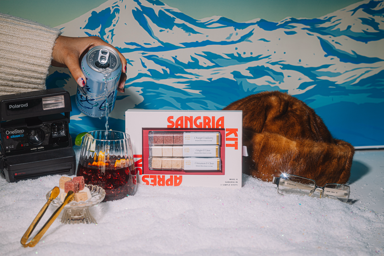 Sangria kit with ingredients and a can of sparking water on snow, against a mountain-themed backdrop with apres ski accessories and a vintage polaroid.