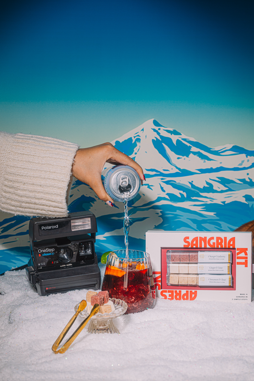 Hand pouring a La croix into a winter sangria. With a mountain and vintage polaroid camera in the background