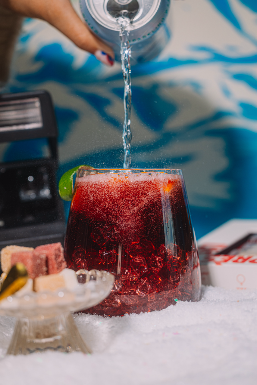 La croix being poured into a sangria wine with ice, garnished with a lime, on a snowy surface, against a blue winter patterned background.