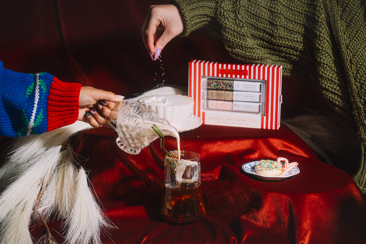Person pouring a drink into a glass on a red tablecloth with a striped box and a small plate of food.