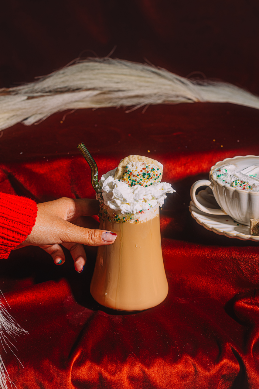 Hand holding a tall glass of iced coffee with whipped cream and sprinkles, on a red surface.