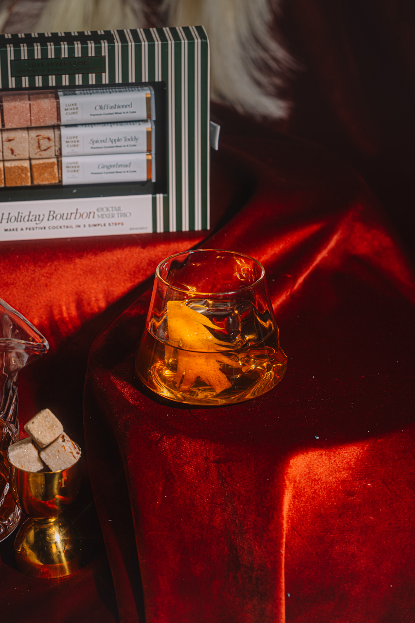 Glass of whiskey with a lemon twist on a red surface, next to a box labeled 'Holiday Bourbon'.