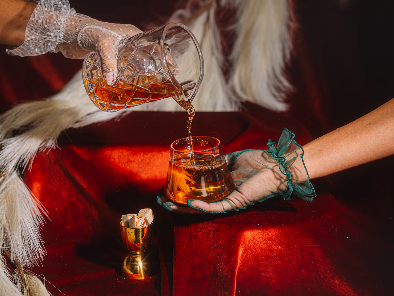 Person pouring whiskey from one glass to another on a red surface with a blurred background