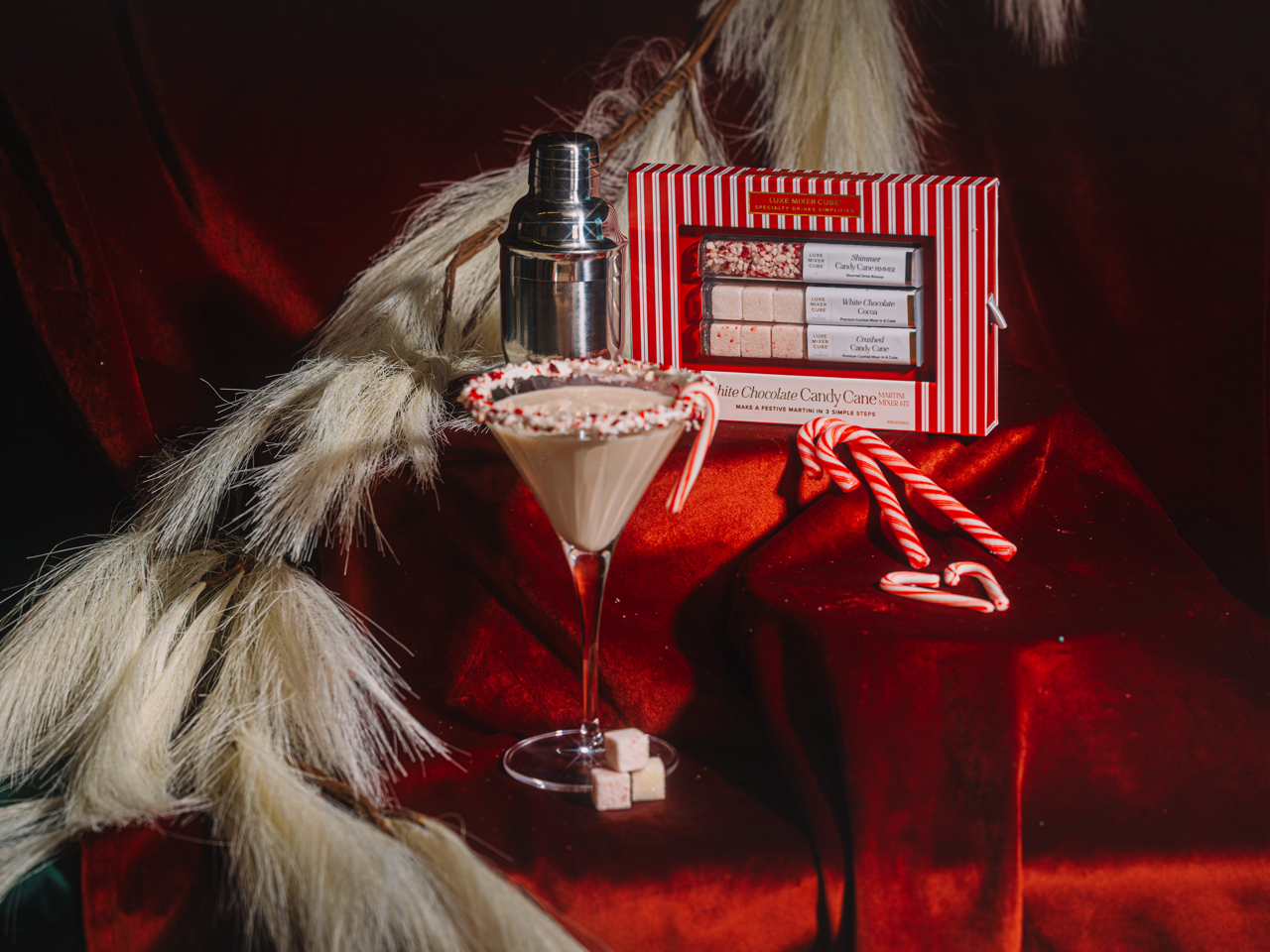 Martini glass with candy cane rim, cocktail shaker, and gift box on a red  velvet surface with fur accents.