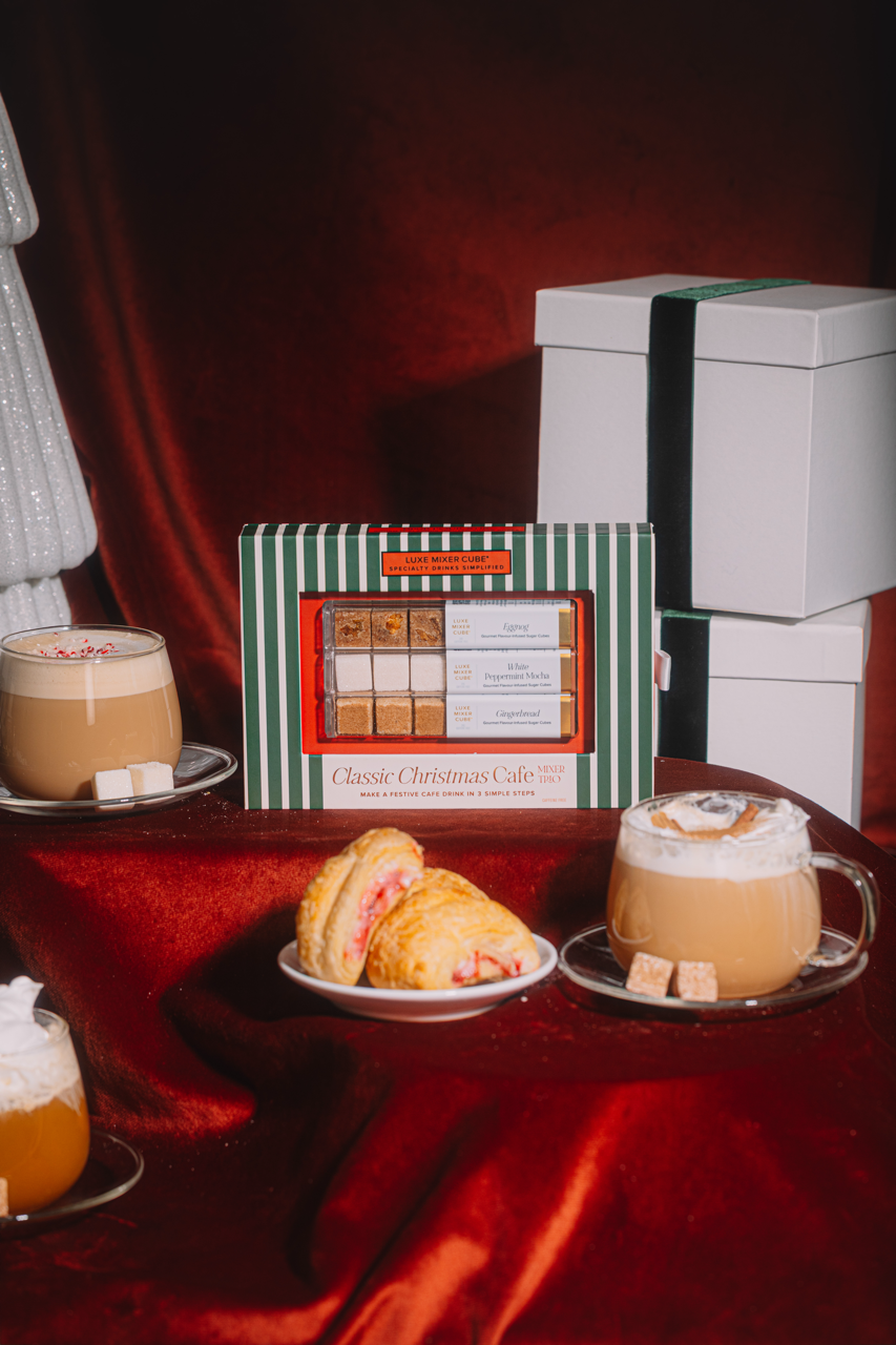 Two cups of coffee with frothy tops, a plate of pastries, and a box of Classic Christmas Cakes on a red fabric background.