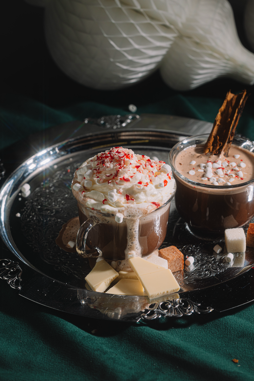 Two hot chocolate drinks with whipped cream and toppings on a silver tray.
