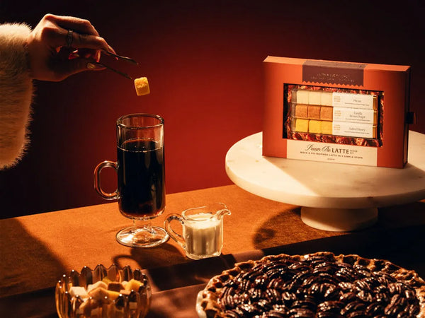 A gourmet Pecan Pie latte kit sits on a white pedestal in a warm, moody setting. A hand wearing a white fur sleeve drops a flavoured sugar cube into a glass of dark coffee.