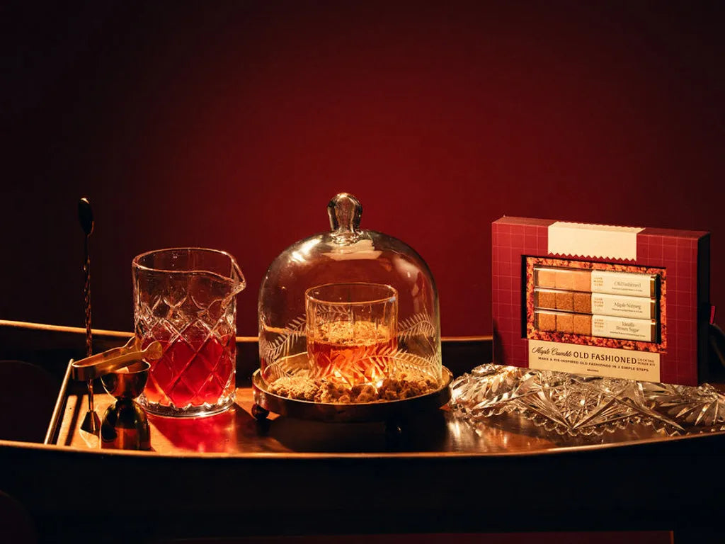 A styled product display featuring a box of Luxe Mixer Cube Maple Crumble Old Fashioned mixer cubes, a crystal mixing glass with red liquid, a crystal decanter, and a glass cloche on a dark wooden tray against a deep red background.