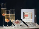 A boxed set of summer champagne desserts displayed beside three champagne flutes with ice and a small glass bowl on a dark studio background. The box reads 'Summer Champagne' and shows assorted pink, cream, and white sweets inside.