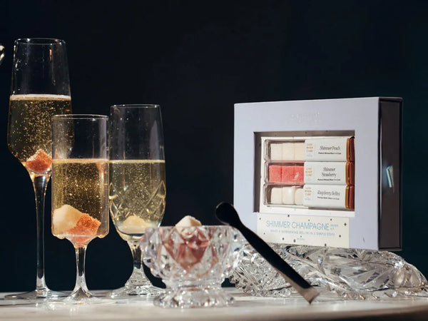 A boxed set of summer champagne desserts displayed beside three champagne flutes with ice and a small glass bowl on a dark studio background. The box reads 'Summer Champagne' and shows assorted pink, cream, and white sweets inside.