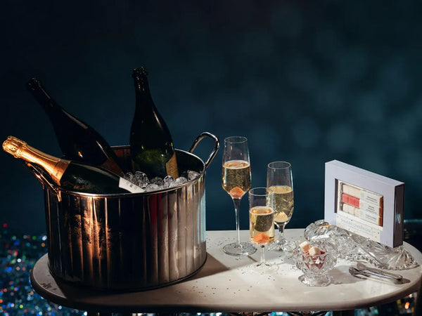 A silver ice bucket holding three champagne bottles, with three filled champagne flutes, a small glass bowl, and a white product box on a round table against a dark blue background.
