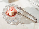 A small clear glass bowl with a faceted scalloped rim holding pink and white sugar cubes, with silver tongs resting beside it on a bright white surface.