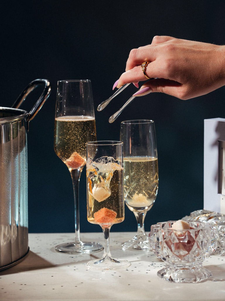 Three champagne flutes filled with sparkling wine on a table, alongside a metal ice bucket, a hand holding tweezers, and a small glass bowl with garnish, set against a dark background.