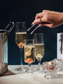 Three champagne flutes filled with sparkling wine on a table, alongside a metal ice bucket, a hand holding tweezers, and a small glass bowl with garnish, set against a dark background.