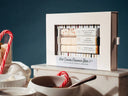 A boxed hot cocoa flavour bar set displayed on a tabletop with bowls, candy canes, and a dark blue background. The white package has a clear window showing several chocolate-flavoured bars inside, and the front label reads 'Hot Cocoa Flavour Bar'.