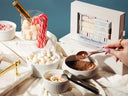 A styled tabletop scene showing a boxed hot cocoa flavour box with several white bowls of ingredients, candy canes, marshmallows, and a hand using a spoon to scoop cocoa powder. The product box is visible on the right against a blue wall.