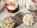 Three glass mugs filled with hot chocolate on a light marble surface, topped with whipped cream and different garnishes: crushed candy canes, white chocolate pieces with colorful sprinkles, and marshmallows. A white fabric napkin is visible in the background.