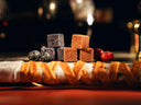 A close-up of assorted square-shaped sweets, including orange-brown and dark purple pieces, arranged on a dark surface with red berries and a white-wrapped item in the foreground against a warm, blurred background.
