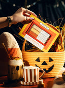 A hand holds a Halloween-themed makeup palette labeled 'Candy Corn Latte' above a woven basket with a jack-o'-lantern face. The scene includes a pumpkin-shaped mug, a striped mug, and autumn decorations in warm, moody lighting.