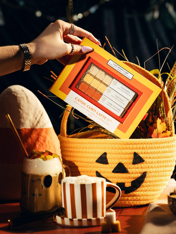 A hand holds a Halloween-themed makeup palette labeled 'Candy Corn Latte' above a woven basket with a jack-o'-lantern face. The scene includes a pumpkin-shaped mug, a striped mug, and autumn decorations in warm, moody lighting.