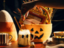 A Halloween-themed tabletop scene with a hand placing candy into a frothy drink in a mug. A woven basket with a jack-o'-lantern face sits behind the drink, alongside a candy corn mug, a bowl of gummy candies, and autumn decorations in warm, low-light lighting.