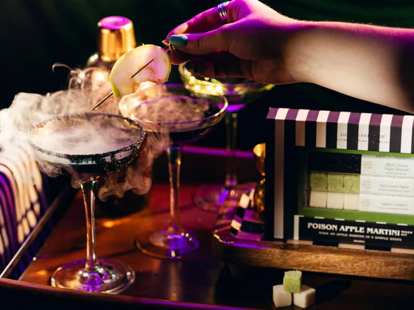 A hand garnishes a cocktail with a sliced apple on a skewer over smoky martini glasses, with a branded box reading 'Poison Apple Martini' and a clapperboard in the background under dramatic purple lighting.