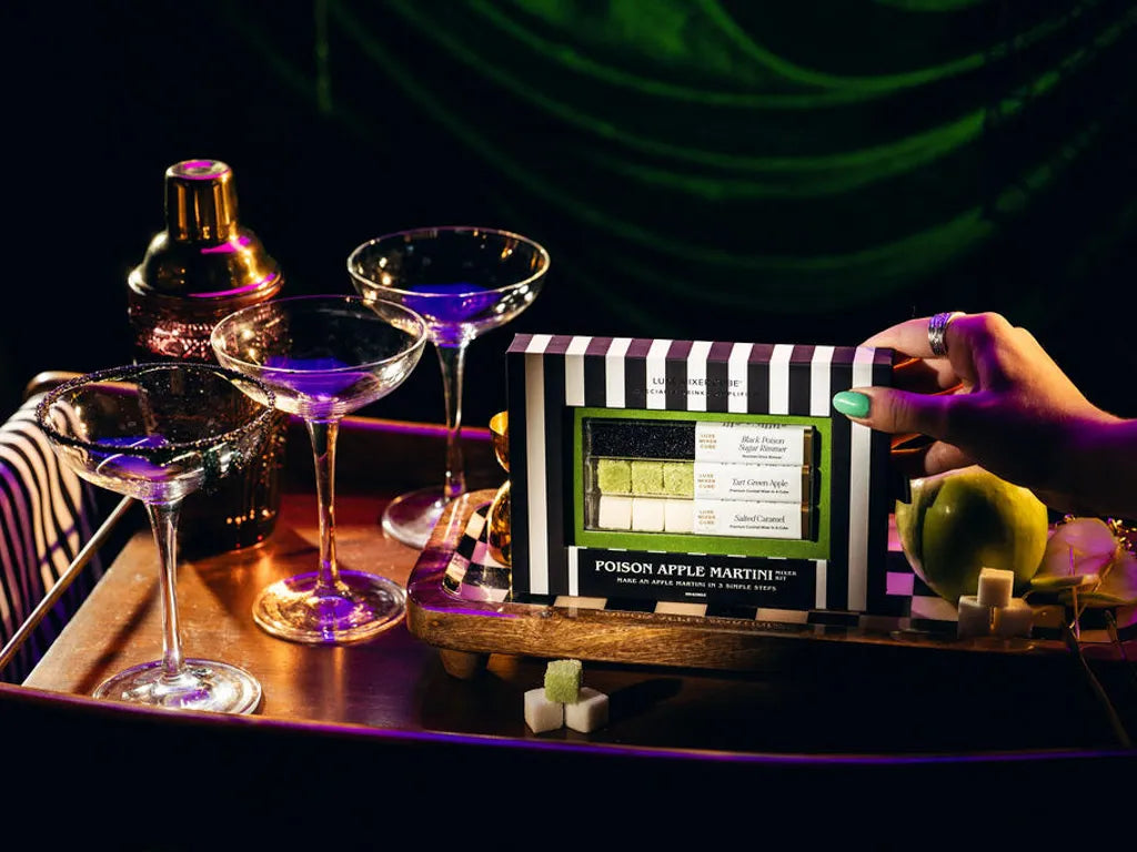 A hand with green nail polish holds a striped box labeled 'Poison Apple Martini' on a tray, surrounded by cocktail glasses, a shaker, and a green apple in a dark, neon-lit setting.