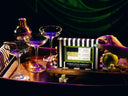 A hand with green nail polish holds a striped box labeled 'Poison Apple Martini' on a tray, surrounded by cocktail glasses, a shaker, and a green apple in a dark, neon-lit setting.