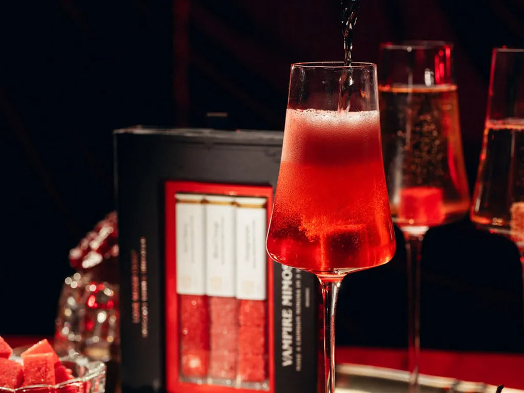 A close-up of a red cocktail in a stemmed glass with a dark liquid being poured into it, set against a dark background with a black and red product box labeled 'VAMPIRE mimosa' and other glasses in the scene.
