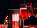 A close-up of a red cocktail in a stemmed glass with a dark liquid being poured into it, set against a dark background with a black and red product box labeled 'VAMPIRE mimosa' and other glasses in the scene.