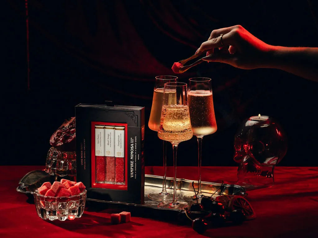 A hand places a red cube into a glass of sparkling champagne on a red tablecloth, next to a box of Vampire Mimosa luxe mixer Cubes. The scene is styled with a skull-shaped candle holder, a small bowl of red cubes, and dark moody lighting.