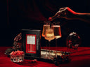 A hand places a red cube into a glass of sparkling champagne on a red tablecloth, next to a box of Vampire Mimosa luxe mixer Cubes. The scene is styled with a skull-shaped candle holder, a small bowl of red cubes, and dark moody lighting.