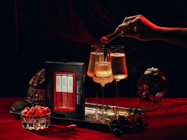 A hand places a red cube into a glass of sparkling champagne on a red tablecloth, next to a box of Vampire Mimosa luxe mixer Cubes. The scene is styled with a skull-shaped candle holder, a small bowl of red cubes, and dark moody lighting.