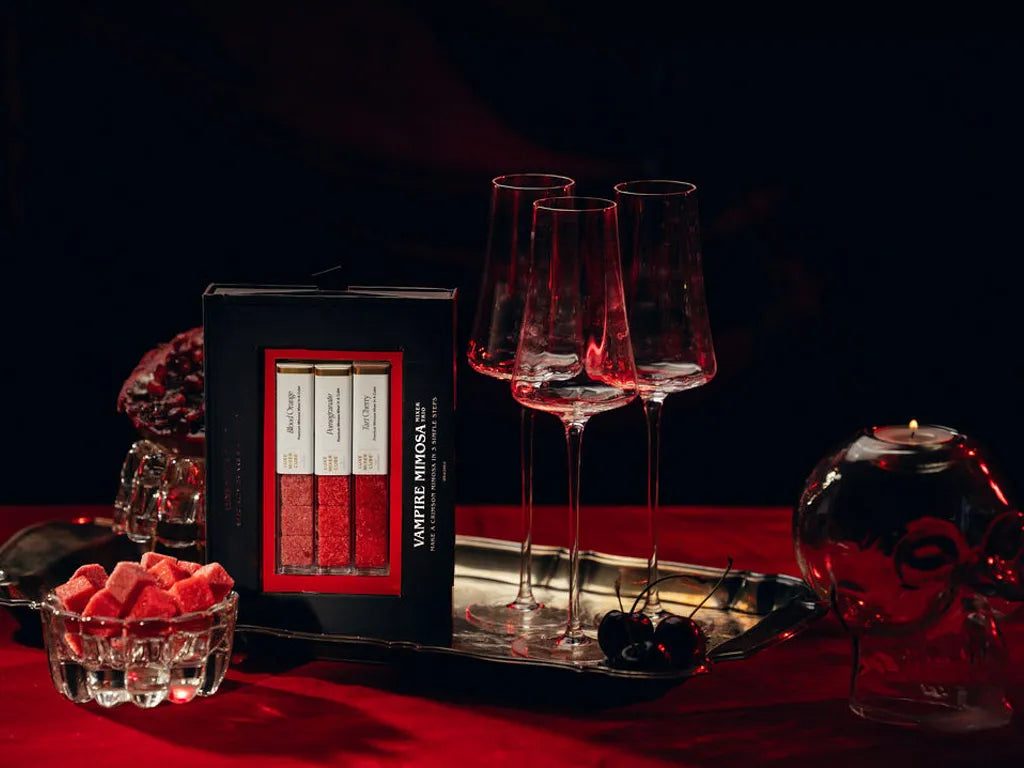 A boxed set of a Vampire Mimosa Mixer trio displayed on a dark tablecloth with three clear wine glasses on a tray, a small bowl of pink cubes, and a skull-shaped glass vessel in a moody red-and-black setting.