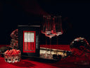 A boxed set of a Vampire Mimosa Mixer trio displayed on a dark tablecloth with three clear wine glasses on a tray, a small bowl of pink cubes, and a skull-shaped glass vessel in a moody red-and-black setting.