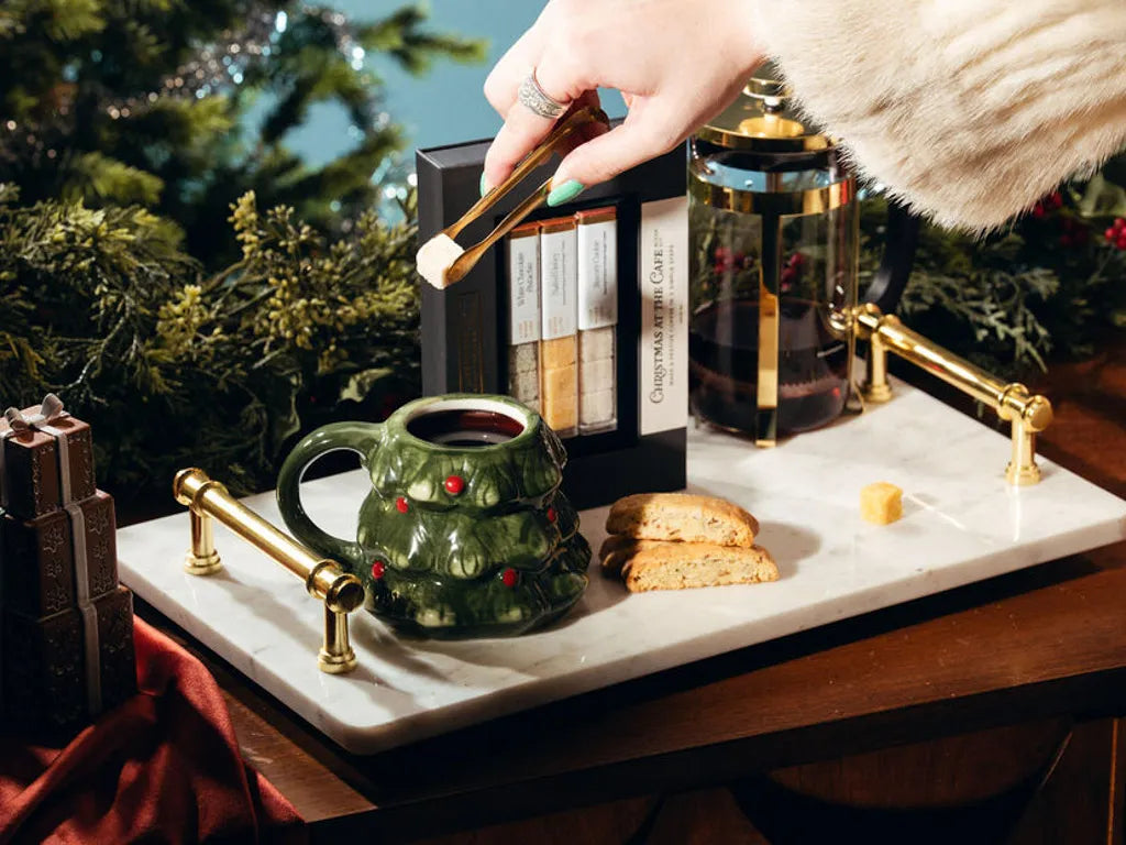 A styled holiday tea and biscotti setting on a white marble tray with gold handles, featuring a green Christmas tree mug, a glass French press, a box of tea, and cookies, with a hand holding a tea bag above.