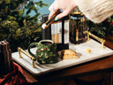 A styled holiday tea and biscotti setting on a white marble tray with gold handles, featuring a green Christmas tree mug, a glass French press, a box of tea, and cookies, with a hand holding a tea bag above.