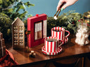 A festive holiday gift set featuring a red gourmet Christmas tea sugar cube kit, two red-and-white striped mugs filled with hot cocoa, sugar cubes in a glass jar, and a decorative wooden house ornament on a wooden table, with a hand placing a sugar cube near the mugs.