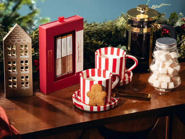 A festive holiday gift set arranged on a wooden table, featuring a red box of gingerbread cookies, two red-and-white striped mugs with saucers, a glass jar of sugar cubes, a black and gold French press, and a decorative gingerbread house ornament.