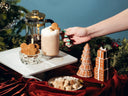A festive holiday beverage setup with a frosted latte topped with a gingerbread cookie, a glass coffee press, cookies in a small bowl, and gingerbread tree and gift decorations on a white platter and red velvet cloth, with a hand reaching in.